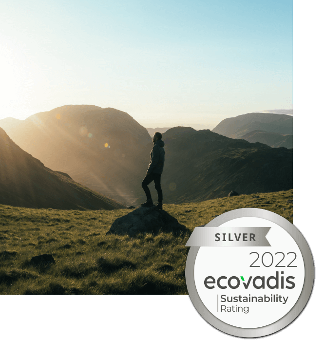A person in the foreground that is standing on a rock in an open field with sun setting. There is a circular icon attached to the bottom right of the image that reads: SIlver 2022 Ecovadis Sustainability Rating.